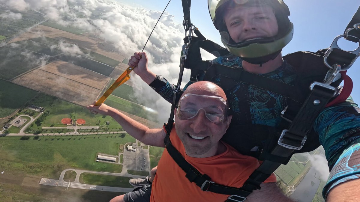 FIG. 03 · March 31, 2025 David grinning mid-air on a tandem skydive above South Florida farmland and clouds, March 31, 2025